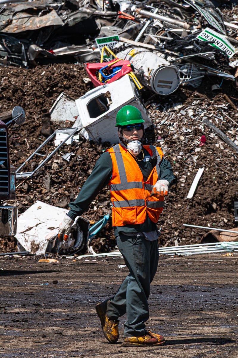 Worker in green hard hat, safety vest, and respirator walking in front of scrap pile filled with appliances and debris.