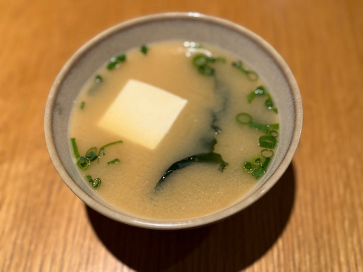 Four images of a traditional Korean and Japanese breakfast spread — grilled mackerel, rice congee with scallions, an assortment of banchan side dishes, and miso soup with tofu.