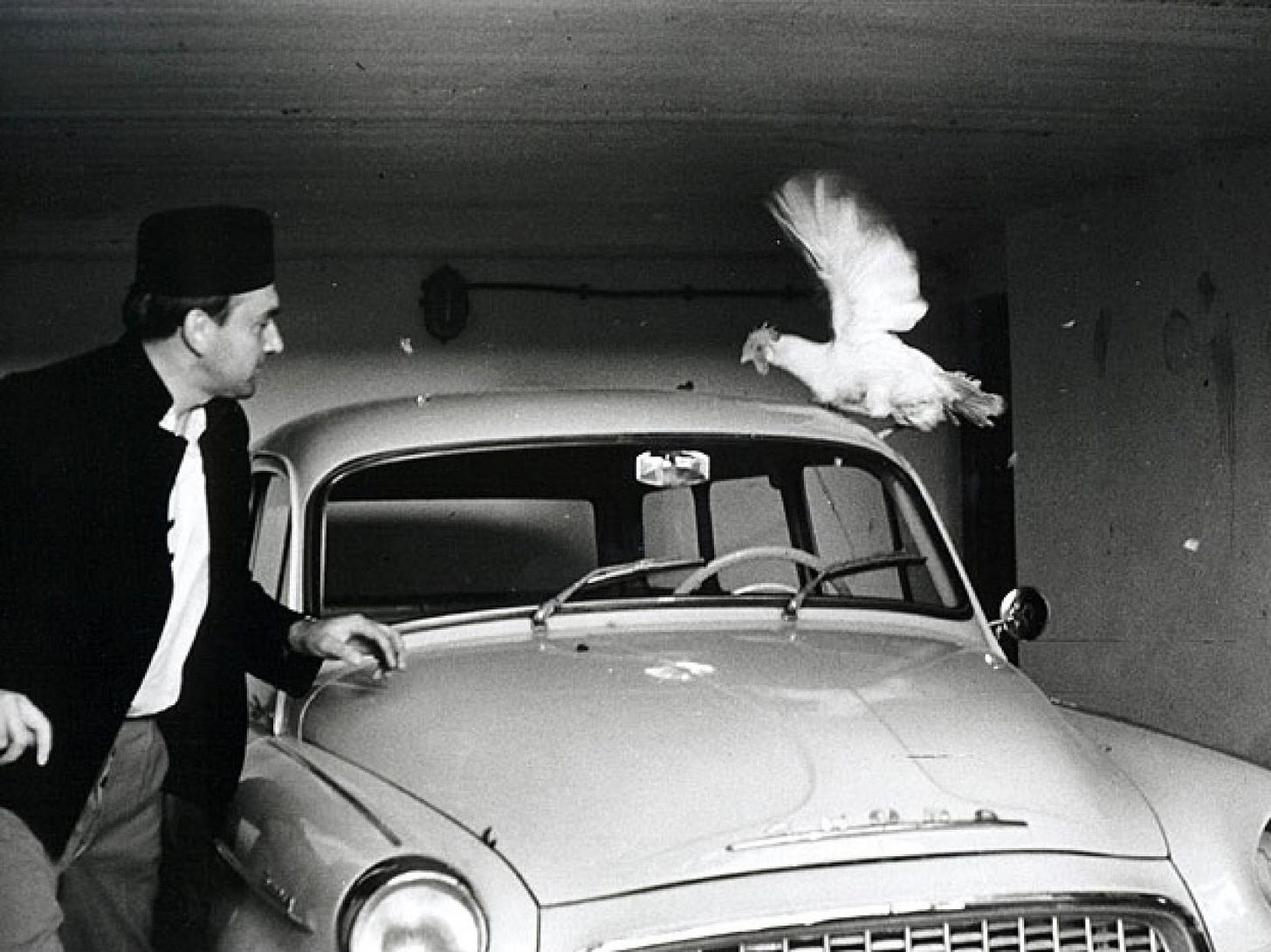 Bambas (Karel Blažek) is confronted by a chicken on the other side of his car.