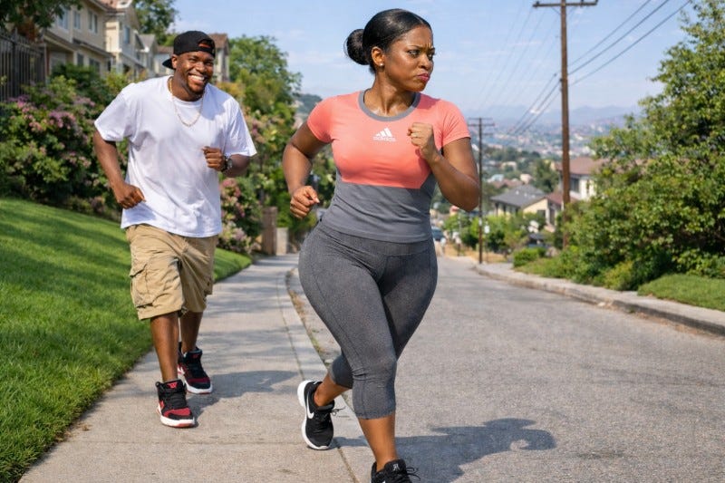 Lady wearing Adidas athletic attire and Nike shoes jogs while a casually dressed man smiles and jogs after her on a residential street.