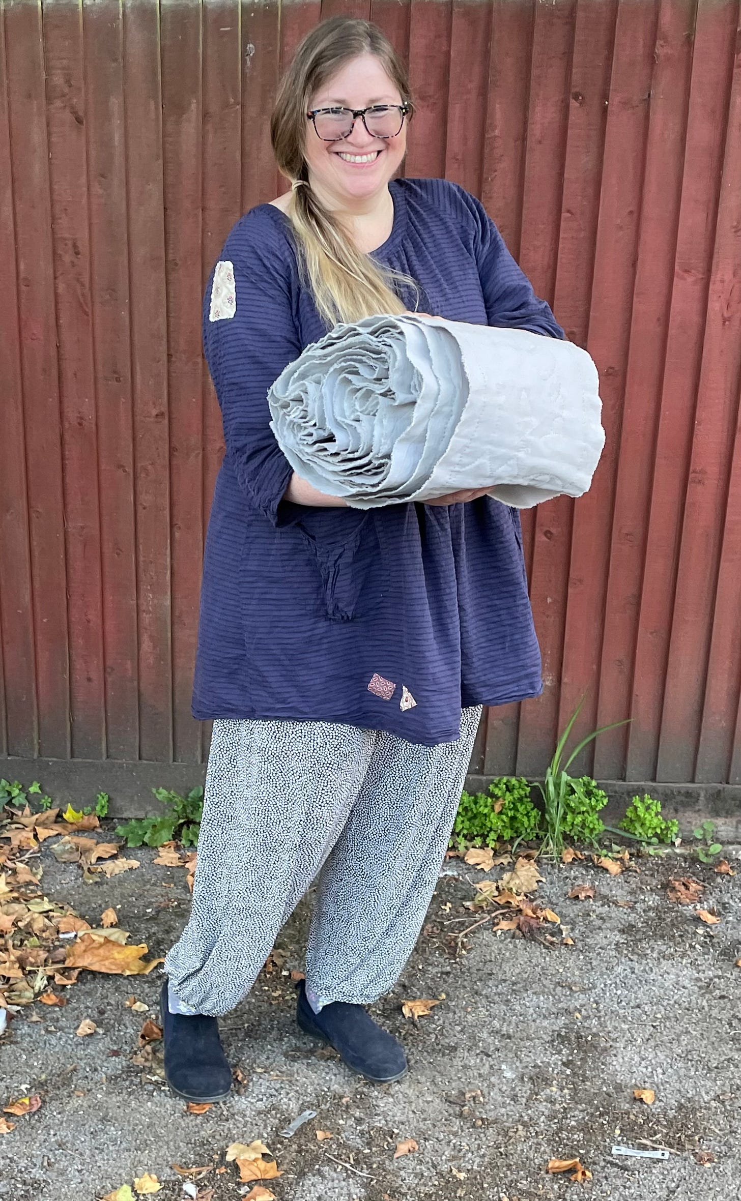 A white woman holding a rolled up piece of quilting, standing beside a wooden fence,