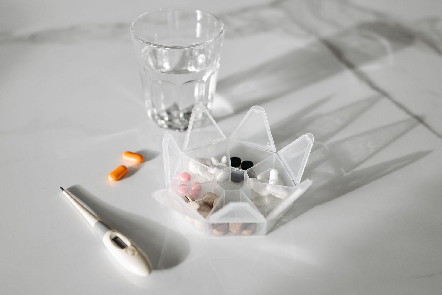 Photo by Pavel Danilyuk on Pexels A pill organizer with medication, a glass of water, and a thermometer on a white surface. (stock photo)