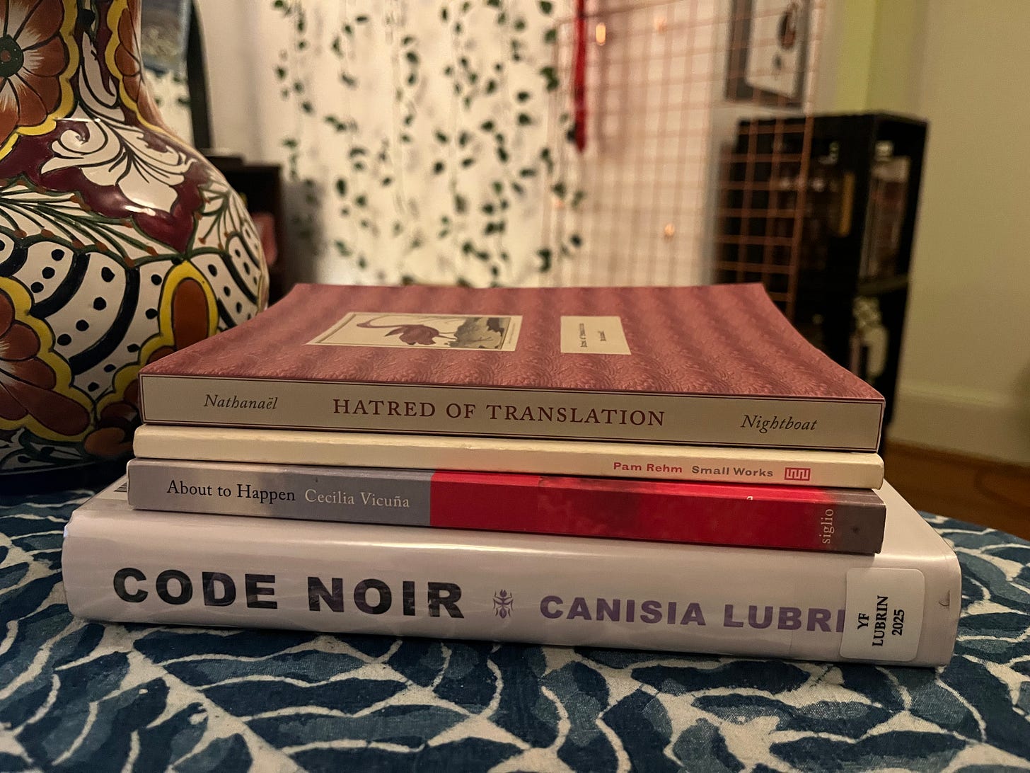 Photo of a stack of four books on a table with a table cloth. Next to them is a decorative jug and in the background a copper rack, fake ivy, and red yarn are (fuzzily) visible. The books' spines are facing towards the viewer. From bottom to top: Code Noir by Canisia Lubrin, About to Happen by Cecilia Vicuña, Small Works by Pam Rehm, and Hatred of Translation by Nathanaël.
