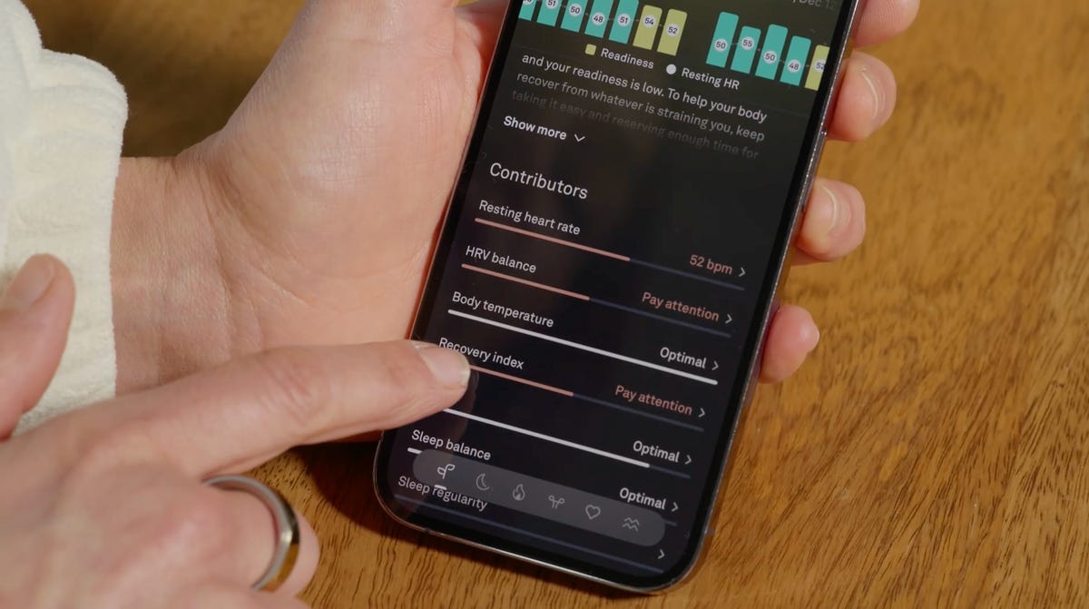 A cellphone is held in a person's hand. The Oura Ring Symptom Radar is pulled up on the screen, listing all the reasons for the user's low readiness score.