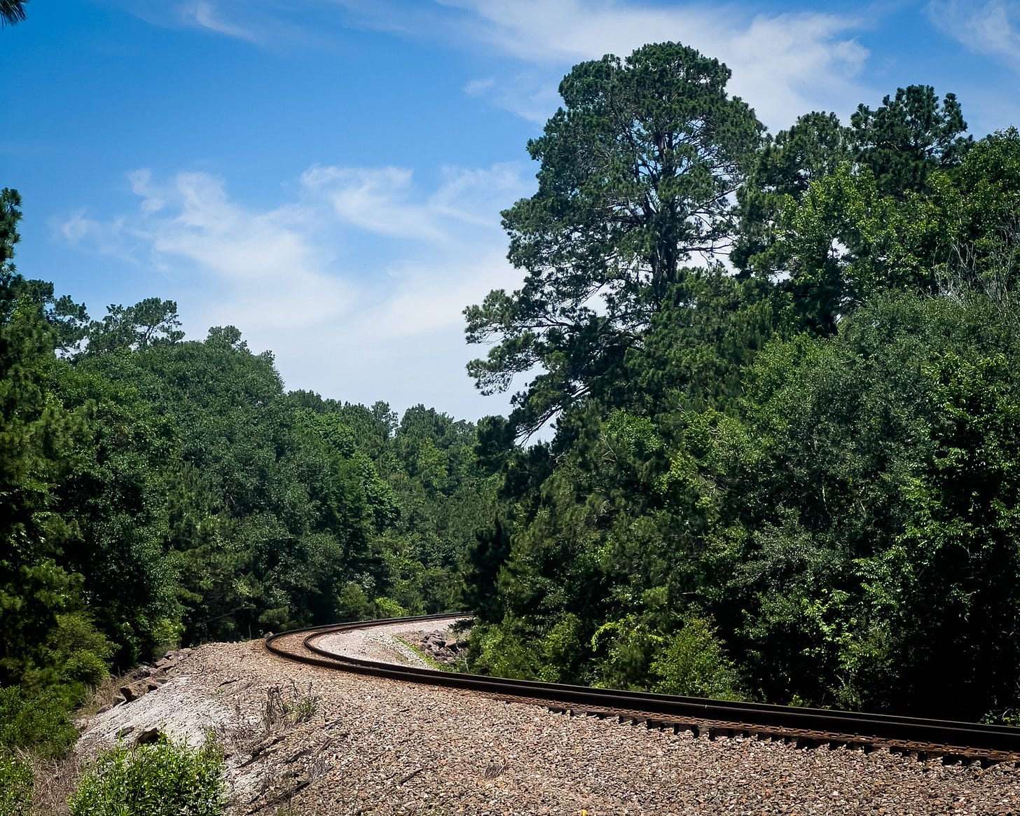 The train track at a bend in the road with trees on either side and a blue sky with white clouds