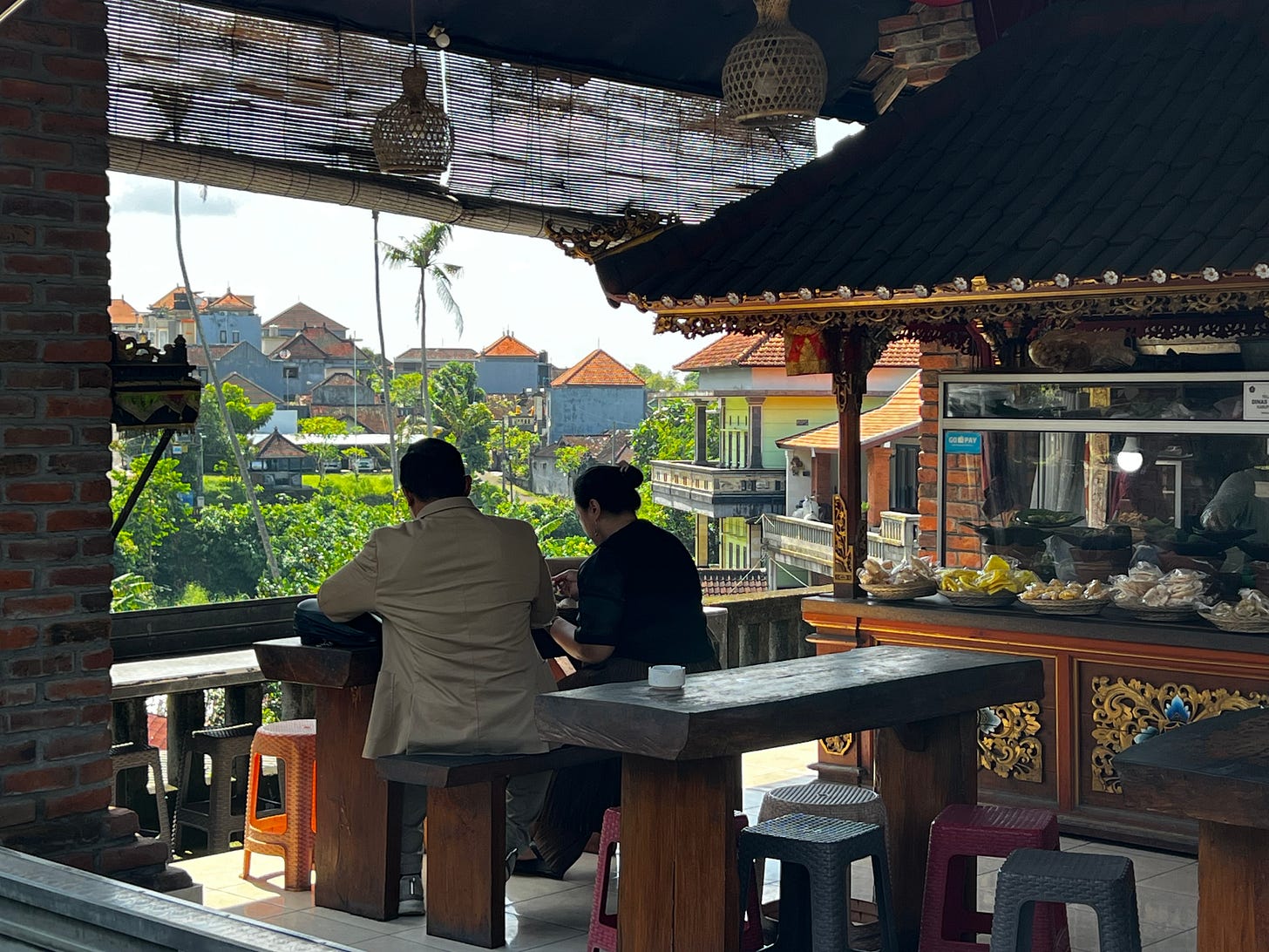 Two people eating at a wooden bench in an open-air warung, looking out over rooftops and palm trees.