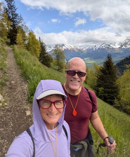 a man and a woman on a steep green hillside with a narrow trail behind them and a rugged mountain range in the distance