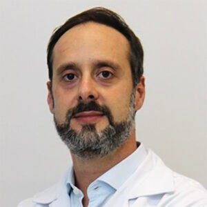 Homem adulto de cabelos curtos escuros e barba grisalha, usando jaleco branco e camisa azul-clara. Olha diretamente para a câmera com expressão tranquila. Fundo liso e claro, em foto de estilo profissional. Homem adulto de cabelos curtos escuros e barba grisalha, usando jaleco branco e camisa azul-clara. Olha diretamente para a câmera com expressão tranquila. Fundo liso e claro, em foto de estilo profissional.