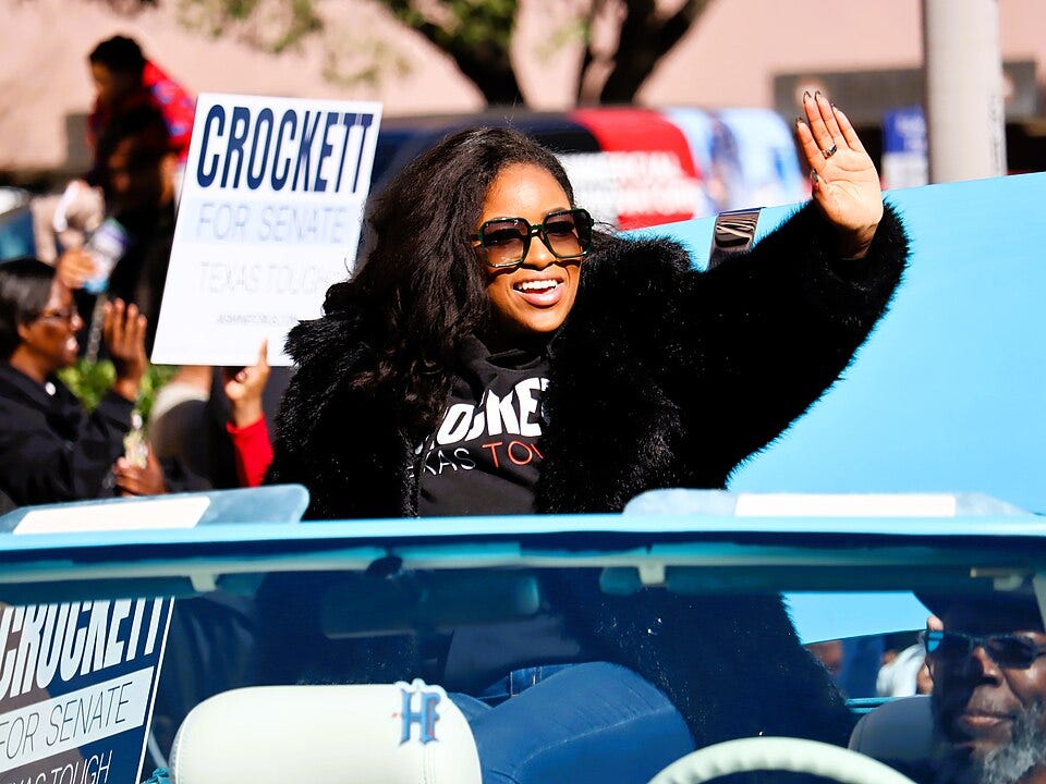 Rep. Jasmine Crockett waves to a crowd from the passenger side of a car.
