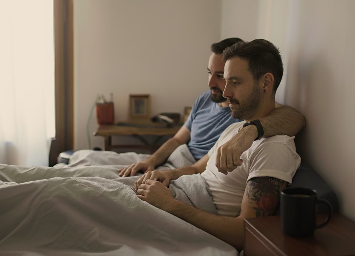 Two men sitting in bed with a coffee mug on the side table.