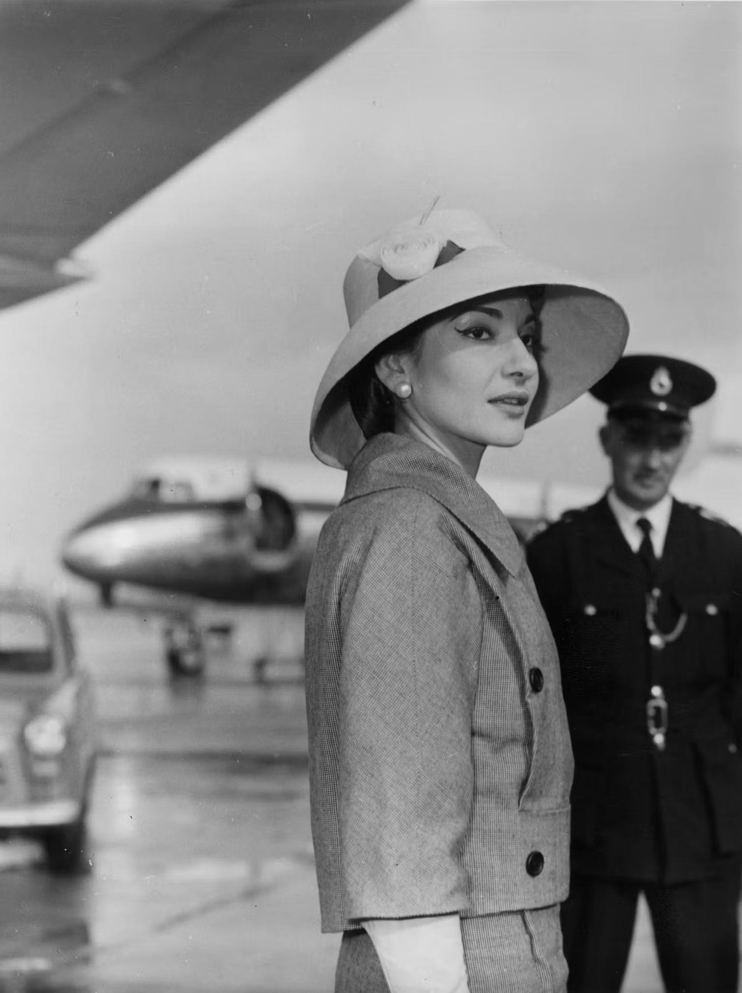 A woman stands almost in profile wearing a broad brimmed hat and feminine day suit on the airport tarmac with a plane in the distance.