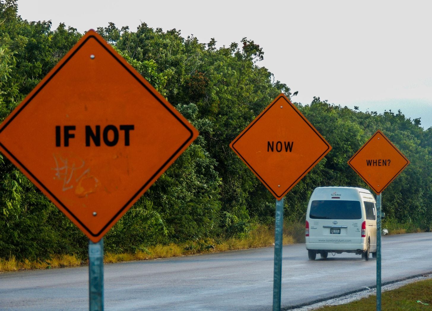 Three orange road signs along a coastal road reading “If not,” “Now,” and “When?” with a van driving away in the background. Tulum Mexico