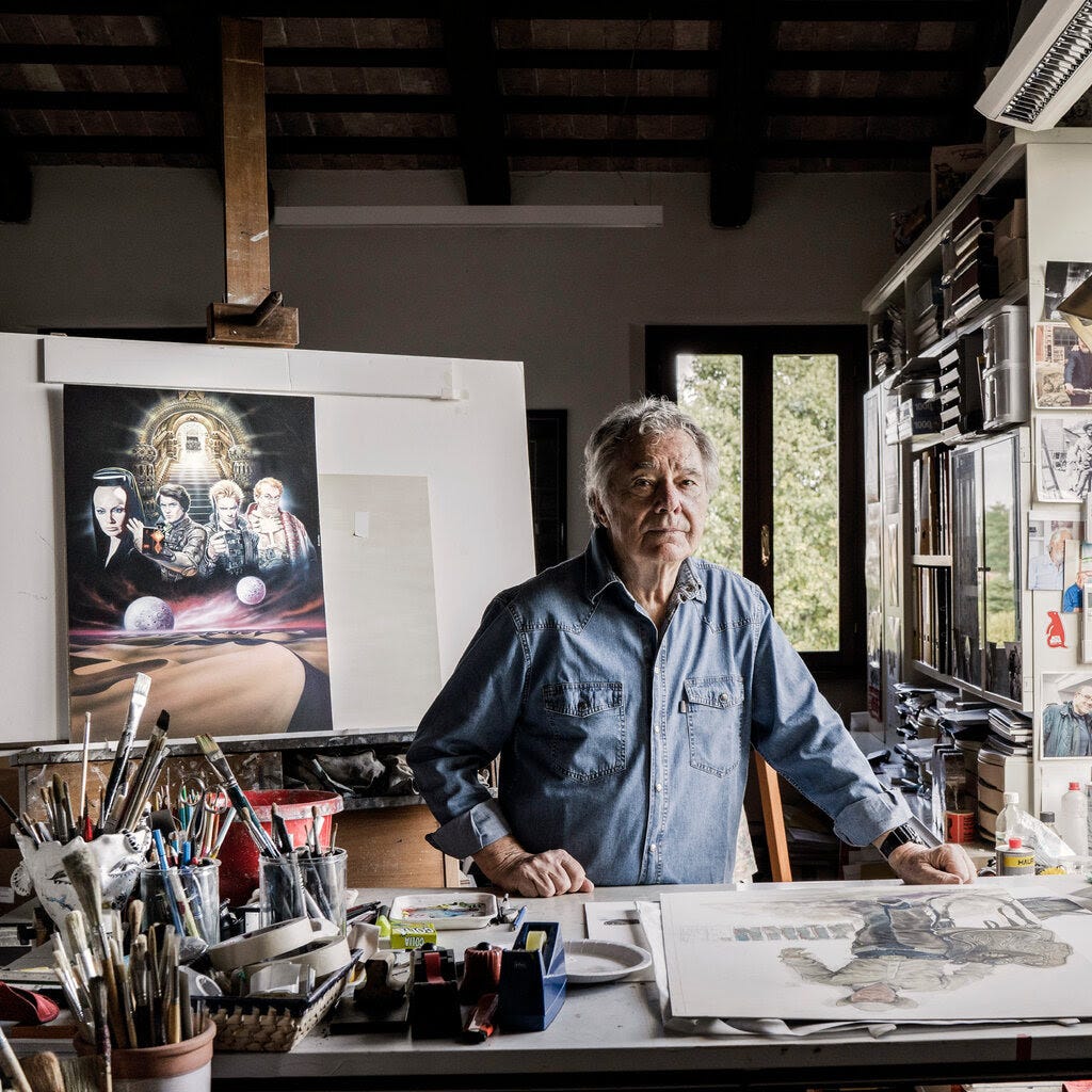 An older Renato Casaro with tousled gray hair stands behind a drawing board surrounded by art supplies.
