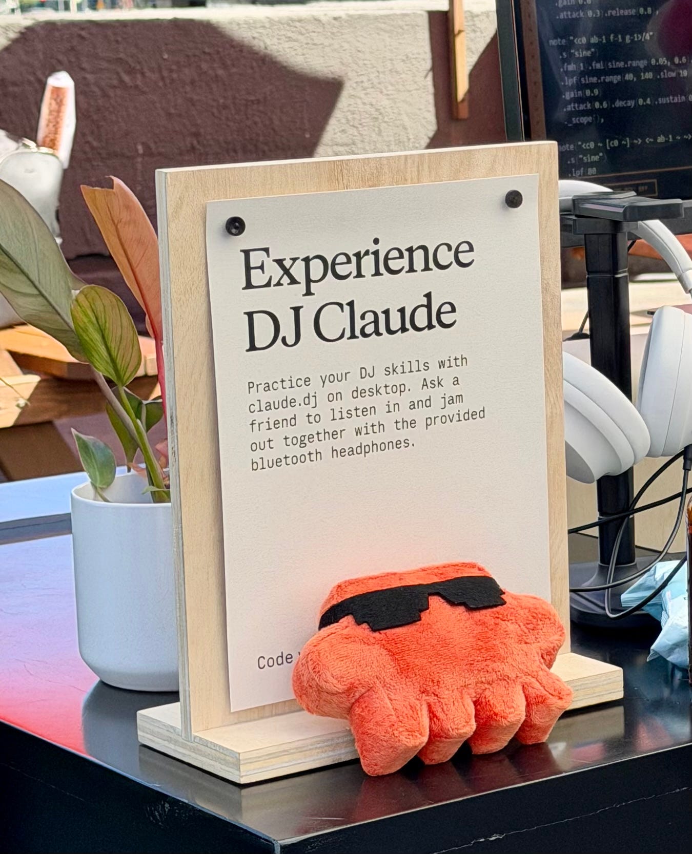 Sign reading “Experience DJ Claude” at Code with Claude 2026 beside headphones, a small plant, and an orange plush toy wearing sunglasses on a demo table. Sign reading “Experience DJ Claude” at Code with Claude 2026 beside headphones, a small plant, and an orange plush toy wearing sunglasses on a demo table.