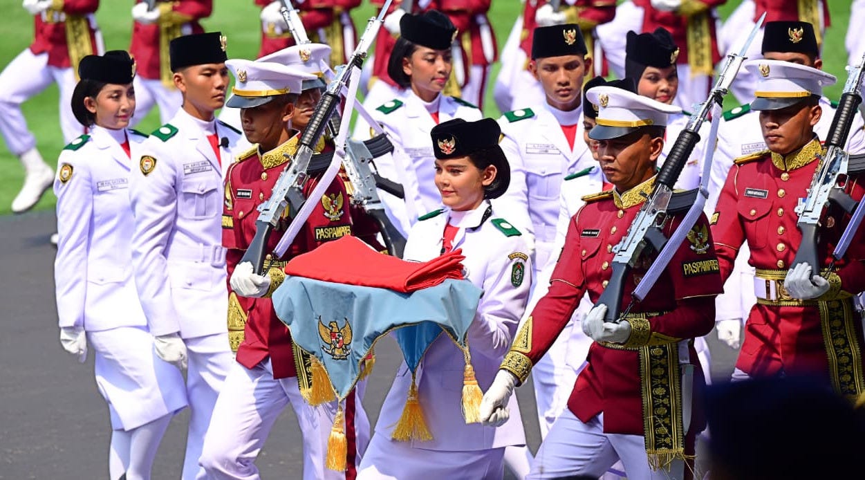 Members of the Indonesian flag-raising troop (Paskibraka) in formal white uniforms, accompanied by honor guards in red ceremonial attire carrying rifles, march in formation while carrying the folded red-and-white national flag on a ceremonial tray during the official 81st Independence Day commemoration in Jakarta. Members of the Indonesian flag-raising troop (Paskibraka) in formal white uniforms, accompanied by honor guards in red ceremonial attire carrying rifles, march in formation while carrying the folded red-and-white national flag on a ceremonial tray during the official 81st Independence Day commemoration in Jakarta.