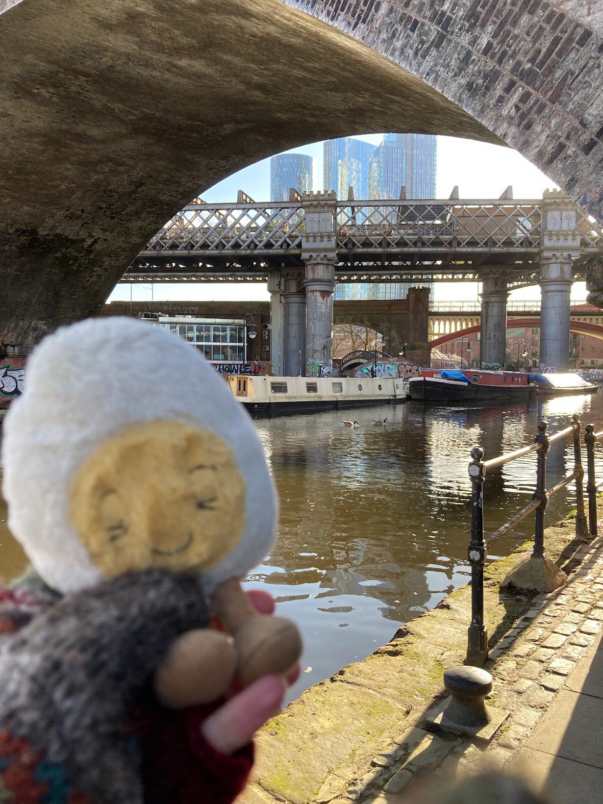 Dippy the Jellycat boiled egg plushie by a Manchester canal, with two canal boats docked and train bridges in the background