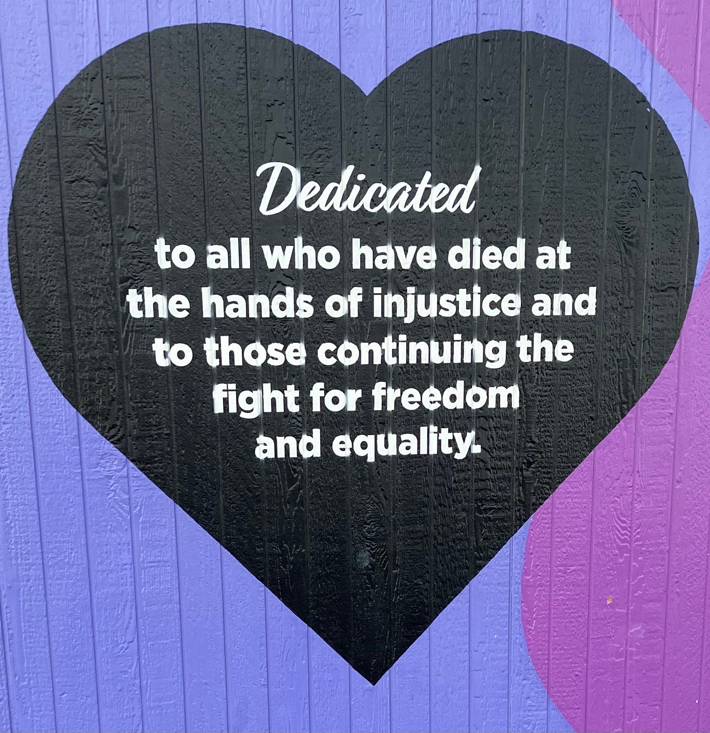 A black heart painted on a blue/purple wall. In white text, it reads " Dedicated to all who have died at the hands of injustice and to those continuing to fight for freedom and equality." A black heart painted on a blue/purple wall. In white text, it reads " Dedicated to all who have died at the hands of injustice and to those continuing to fight for freedom and equality."