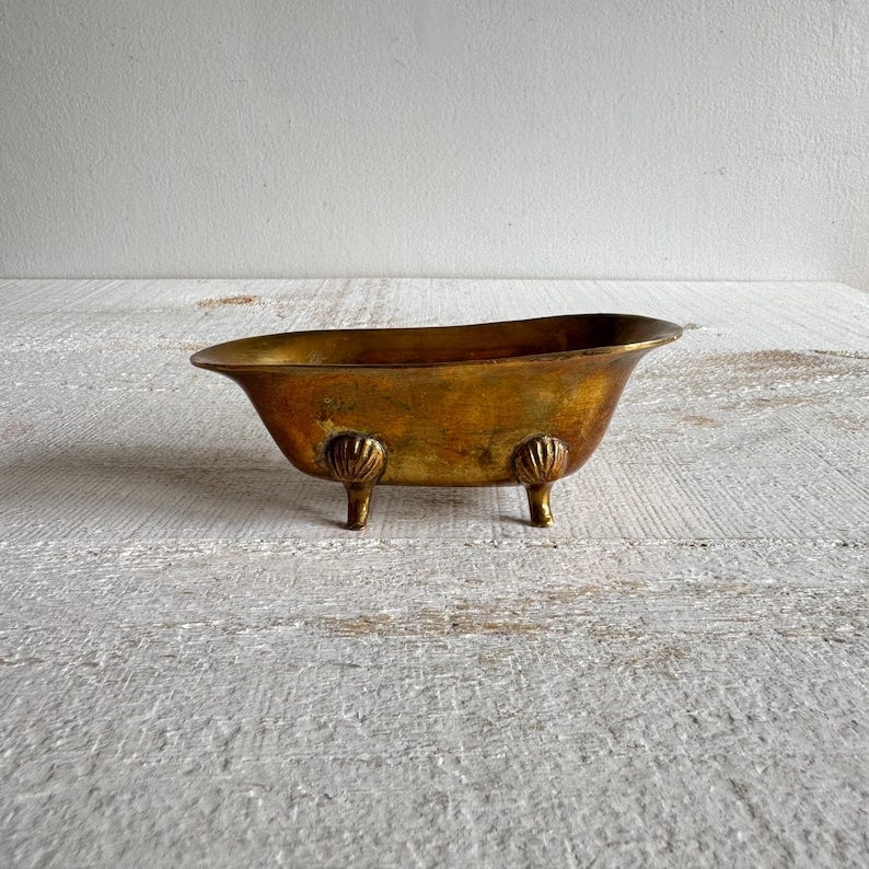 May include: A small, gold-colored, metal bathtub with four feet. The bathtub is on a white, wooden surface.