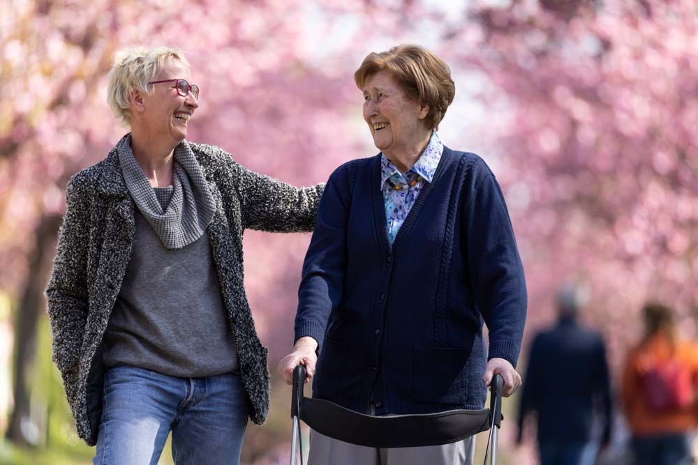 Image of two elder women enjoying a walk through a cherry blossom orchard, one woman is using a walker.