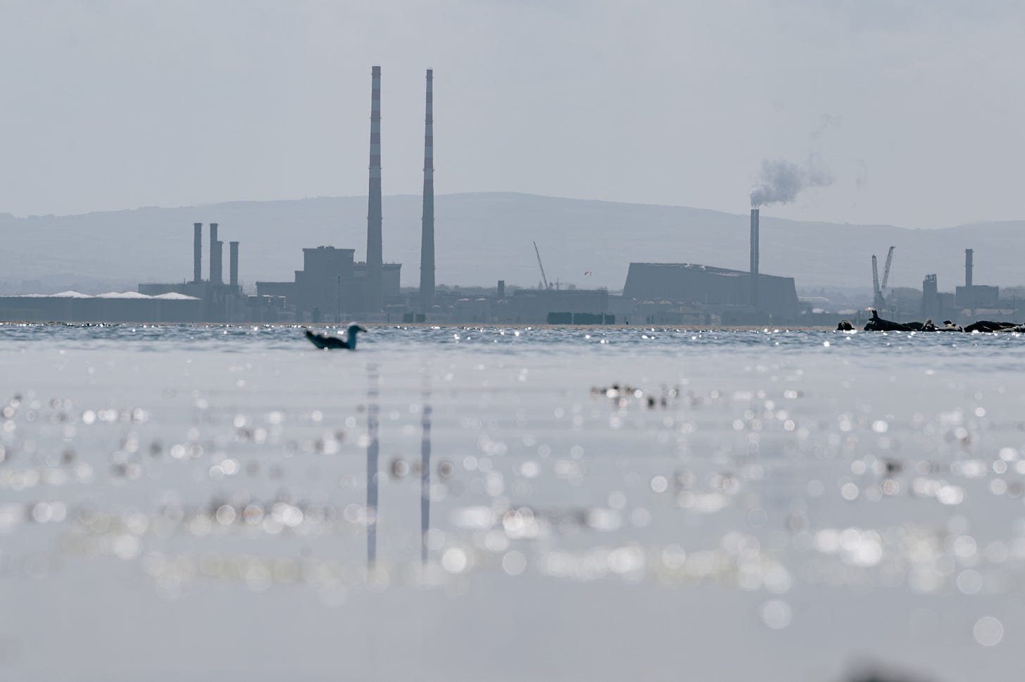 The two red and white chimneys of the Poolbeg towers seen across wet sand at low tide, a small boat on the water and low hills along the horizon. The two red and white chimneys of the Poolbeg towers seen across wet sand at low tide, a small boat on the water and low hills along the horizon.