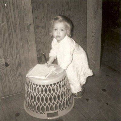 Girl, about age 2, stands in robe and slippers to write on a notepad on an upturned laundry basket