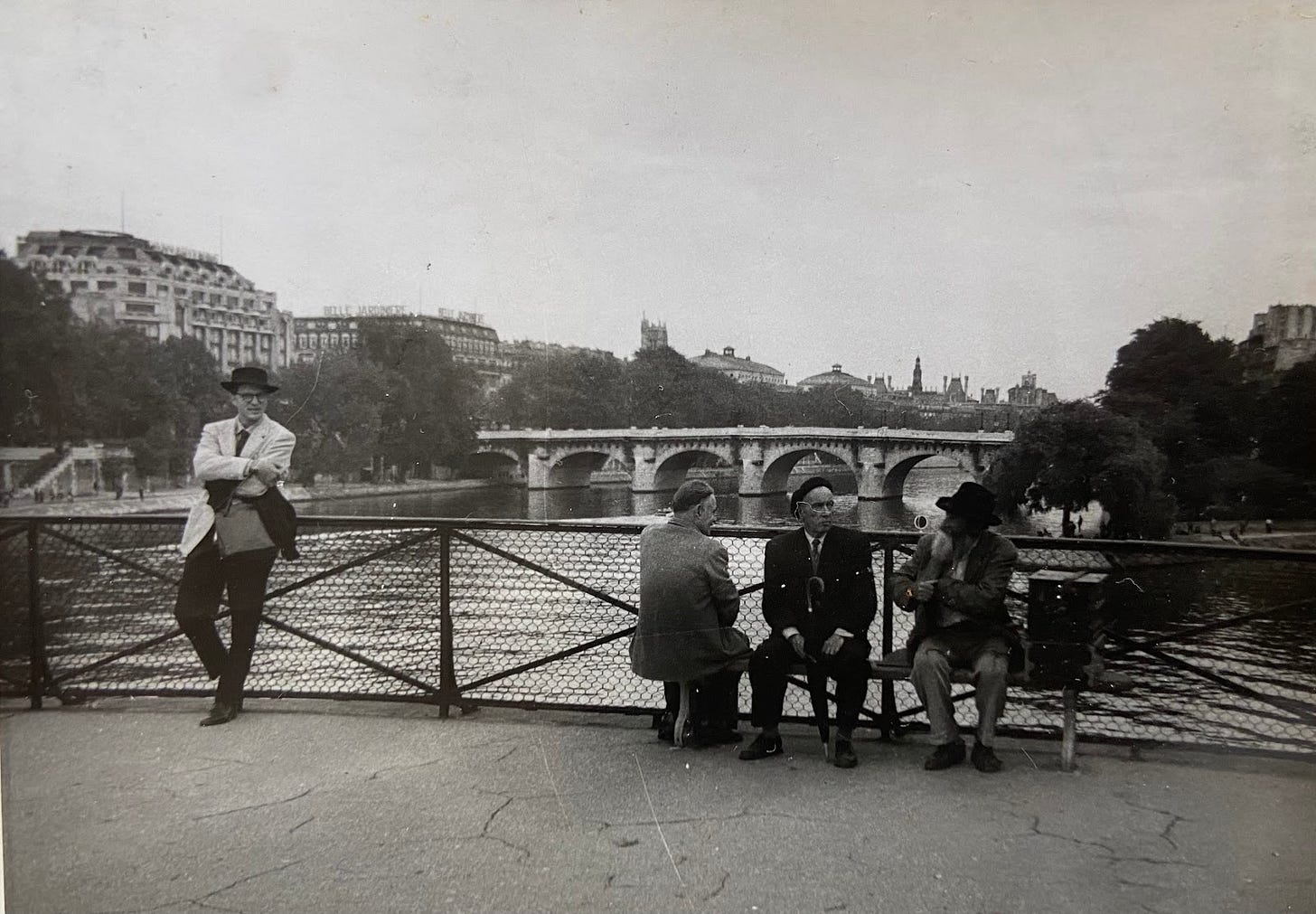 An elegant man wearing a fedora and sport coat with arms crossed leans against the railing on a bridge over a river in Paris and looks toward a group of older men wearing berets seated on a bench.