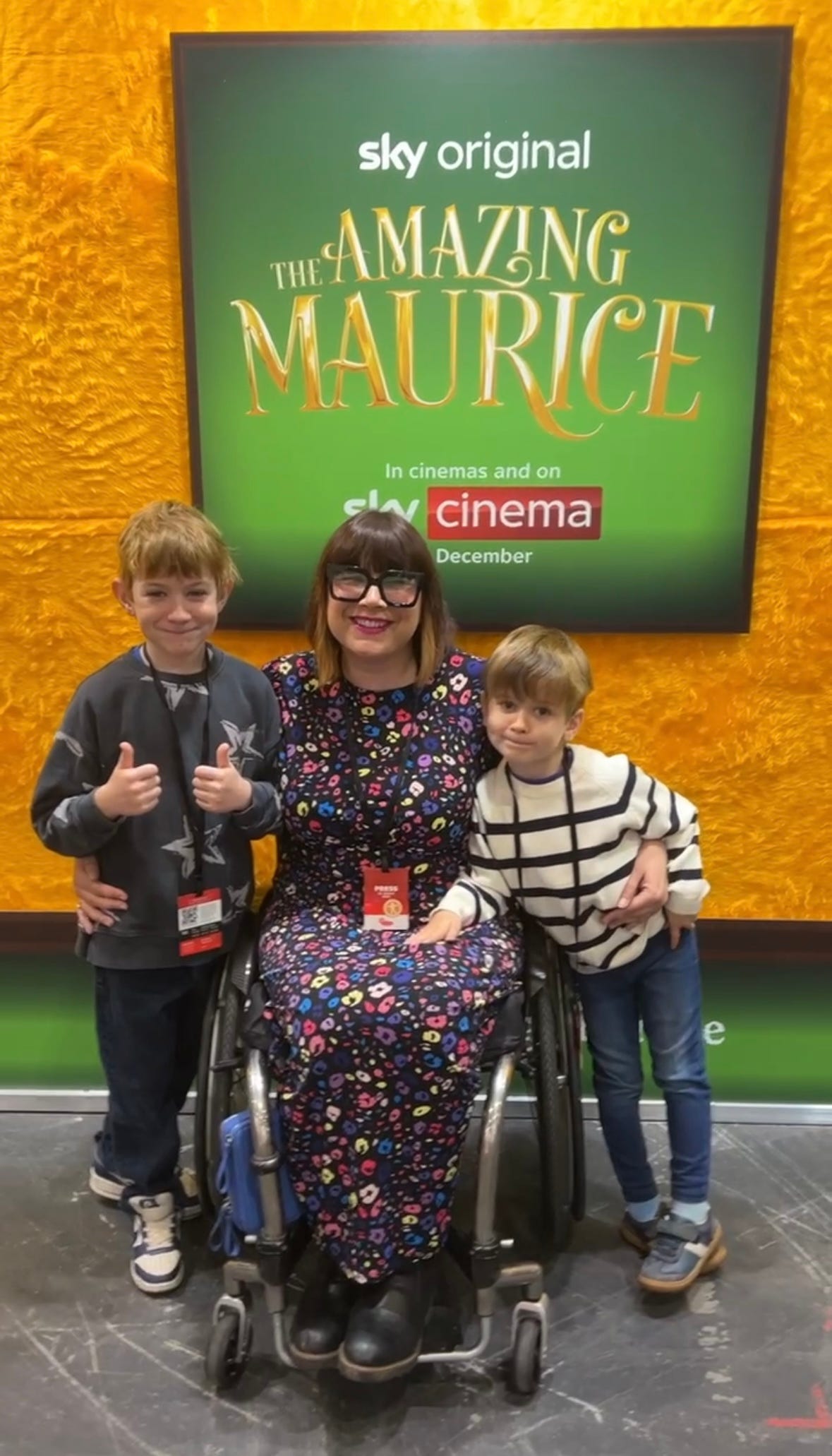 Nina a middle aged white wheelchair user with brown bobbed hair and a fringe. Is wearing a rainbow leopard print maxi dress and smiling with her two small kiddos either side of her. Infront of a movie poster for a cartoon cat