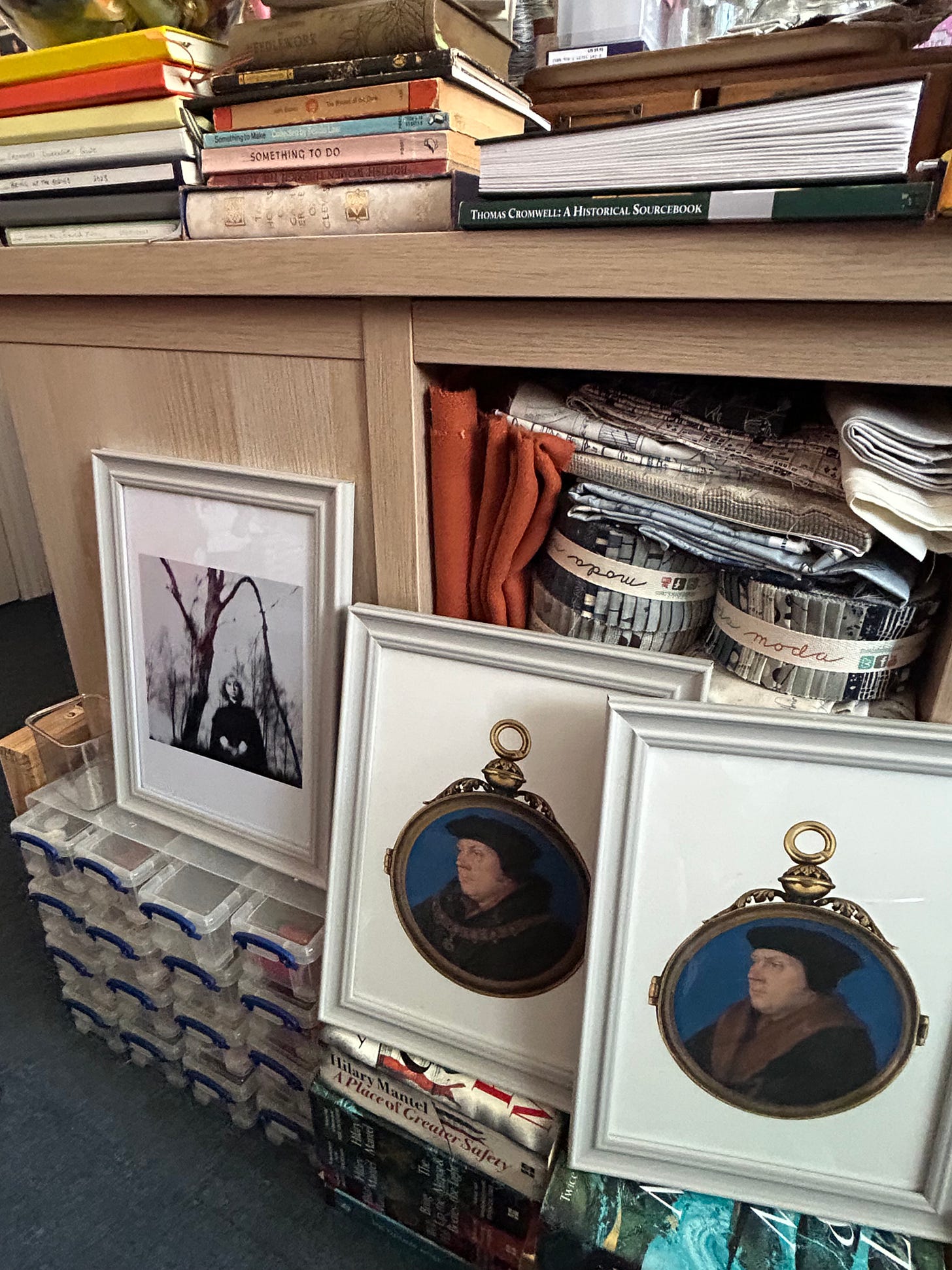 Portraits of Thomas Cromwell and Hilary Mantel rest on books by Mantel. A shelf in the background contains fabric,and there are books on the top. 