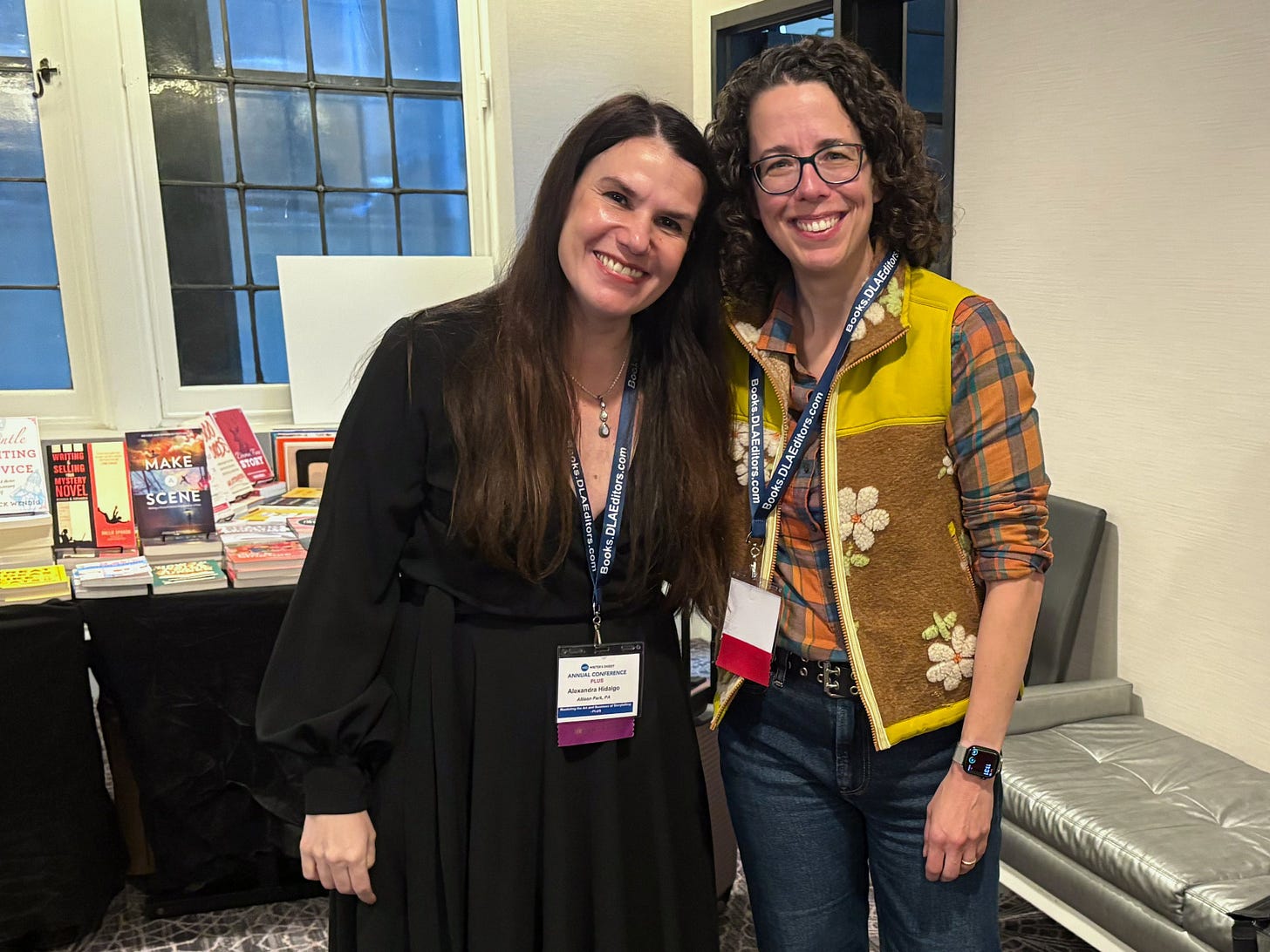 Two women (Alexandra Hidalgo and Jane Friedman) stand together in a hotel lobby. They wear conference lanyards and smile at the camera.  There is a table featuring books for sale behind them. 