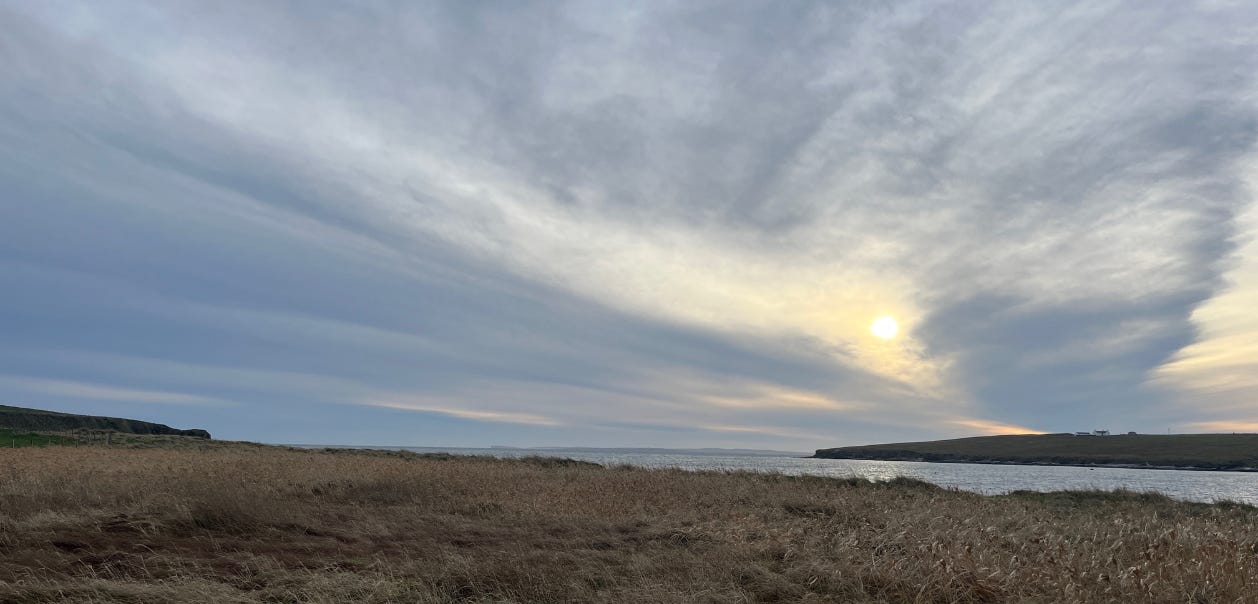 a moody photo of Hoy, Orkney. Grey skies, the sun is jus peeping through and a sweep of light grass with the sea in the background