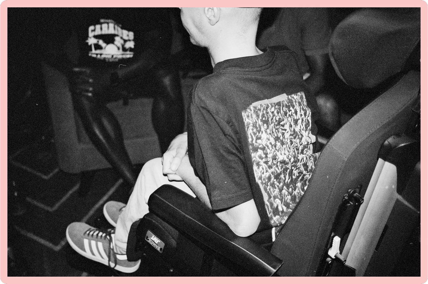 A black-and-white photo, taken from slightly behind, of a young white man who is a wheelchair-user. His hands are crossed on his lap, as if he is listening to someone off-camera, but his face is not visible.