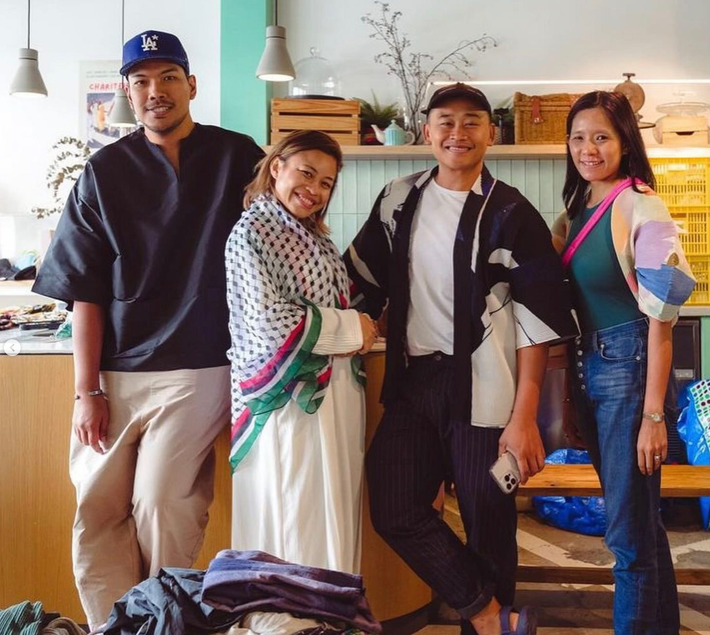 Four people are standing in a brightly lit indoor space, smiling at the camera. The person on the far left is wearing a black shirt, beige pants, and a blue baseball cap. Next to them is a person in a white dress with a colorful scarf, followed by a person in a white t-shirt with a black and white jacket and dark pants. The person on the far right is wearing a colorful jacket, blue jeans, and a green top. The background includes a counter with various items and a shelf with plants and decorations. Four people are standing in a brightly lit indoor space, smiling at the camera. The person on the far left is wearing a black shirt, beige pants, and a blue baseball cap. Next to them is a person in a white dress with a colorful scarf, followed by a person in a white t-shirt with a black and white jacket and dark pants. The person on the far right is wearing a colorful jacket, blue jeans, and a green top. The background includes a counter with various items and a shelf with plants and decorations.