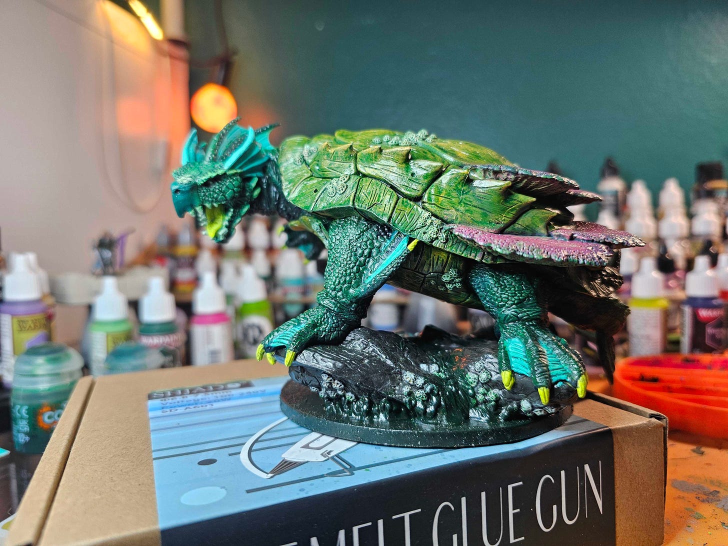 A large turtle figurine stands with it's mouth open to snap and it's foot poised to swipe an unwary hero. It stands perched on barnacle-covered rocks. The figurine rests on a  hot glue gun box and behind it are many small bottles of paint.