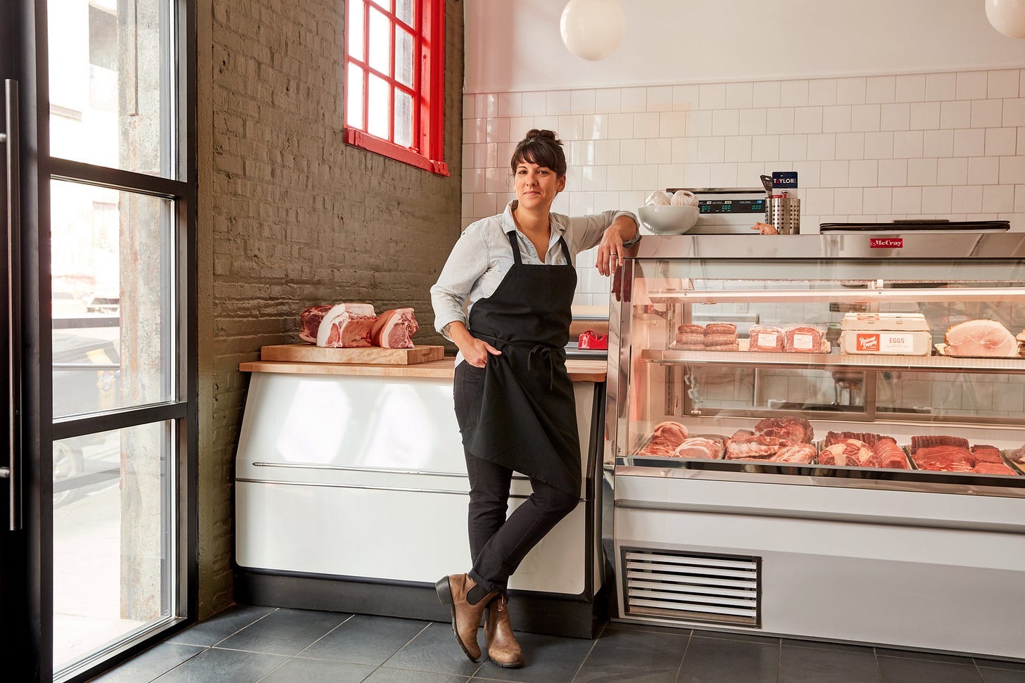 A woman with brown hair, swept up in a bun, wearing a black apron, leans casually against a case featuring cuts of meat. 