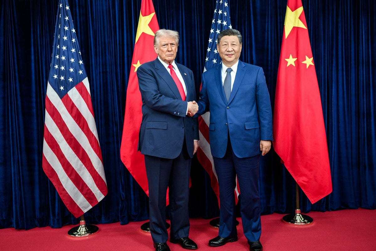 Two men in dark suits shake hands on a red carpet. The man on the left wears a red tie and stands in front of American flags. The man on the right wears a blue tie and stands in front of Chinese flags. Blue curtains form the background with gold stands holding the flags. Two men in dark suits shake hands on a red carpet. The man on the left wears a red tie and stands in front of American flags. The man on the right wears a blue tie and stands in front of Chinese flags. Blue curtains form the background with gold stands holding the flags.