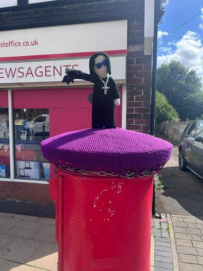 A knitted figure of Ozzy Osbourne perched atop a Royal Mail post box, complete with long black hair, dark sunglasses, and what appears to be a cross necklace. The Prince of Darkness himself is rendered in black wool, sitting on a vibrant purple knitted cover that's been lovingly yarn-bombed onto the red pillar box. His right arm is extended, holding a bat (which is also made of black wool) at arm's length.
Behind Oxzy, a newsagent's shop provides a decidedly mundane backdrop to this delightfully bonkers tribute to the Birmingham metal legend.