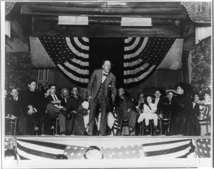 Photo shows Eugene V. Debs making a speech from a stage on which several other men, women, and a young girl are seated behind him; also shows 48-star American flags hanging at the back of the stage and along the front as bunting.