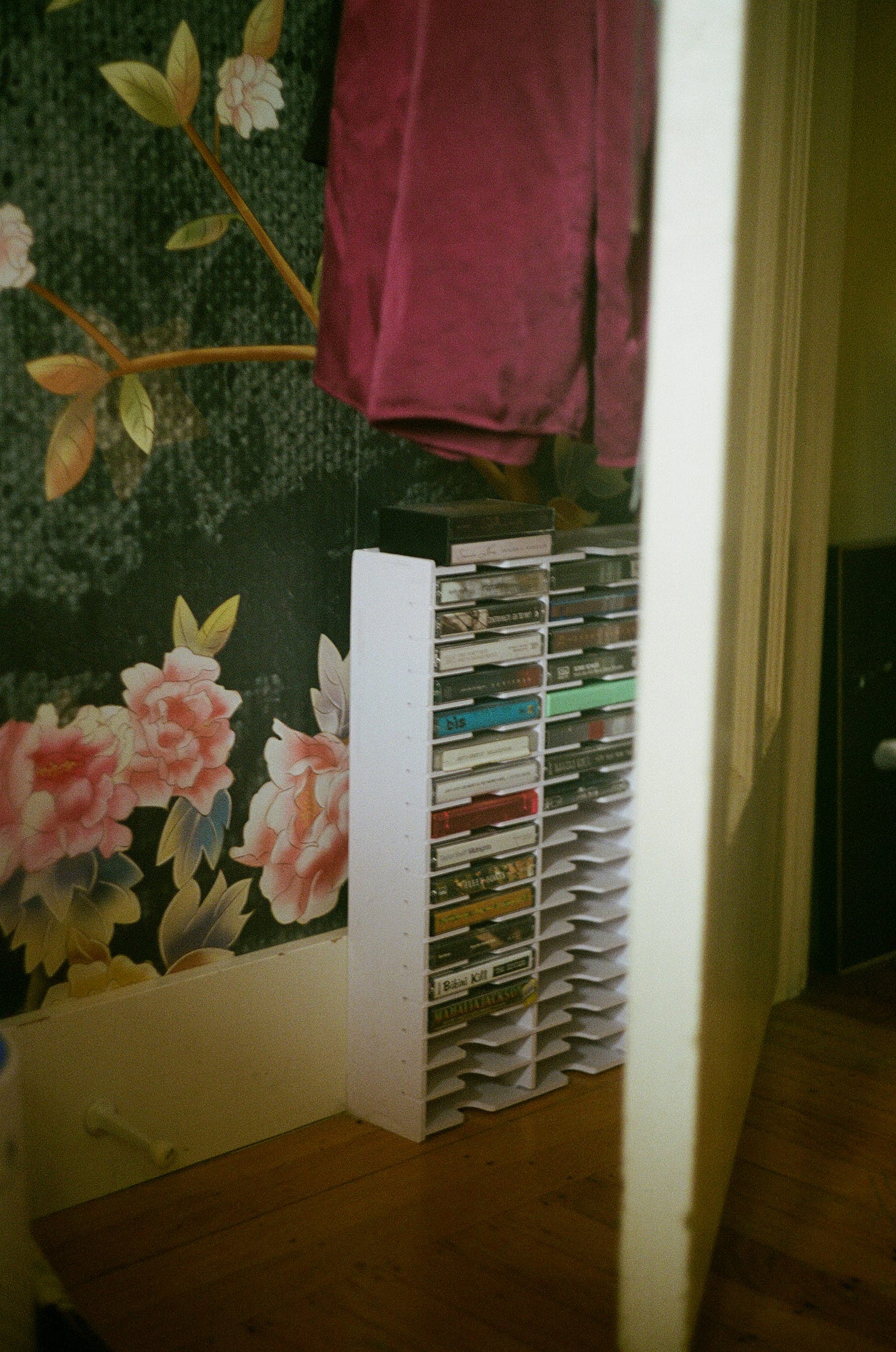 A white cassette tape rack filled with tapes stands against a floral-patterned wall. A burgundy garment hangs above, partially covering the view.