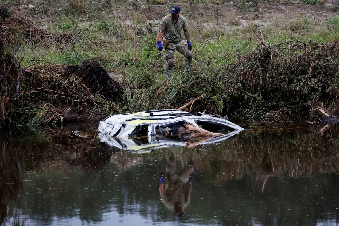 A person on a river's bank surveys a car that's partially submerged in the war.