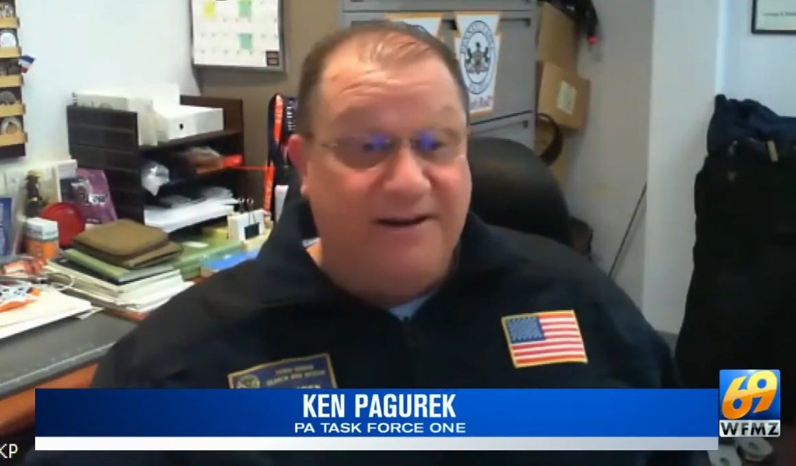 In an office, in front of a desk piled high with books and papers, a balding burly rescue chief, wearing glasses and a navy blue uniform shirt, sits for a tv interview. His face is animated as he talks about how well a rescue dog performed. In an office, in front of a desk piled high with books and papers, a balding burly rescue chief, wearing glasses and a navy blue uniform shirt, sits for a tv interview. His face is animated as he talks about how well a rescue dog performed.