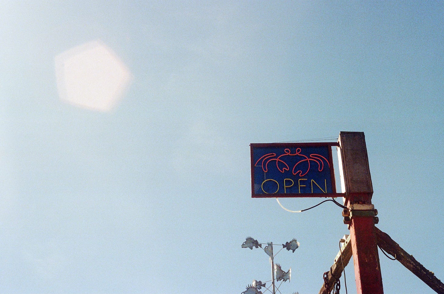 A film photograph of a mostly blue sky, with a light leak in a pentagon shape hovering in the upper lefthand corner. Rising into the frame on the right is a beam with a neon sign on top. The sign reads, "OPEN," with the image of a crab.