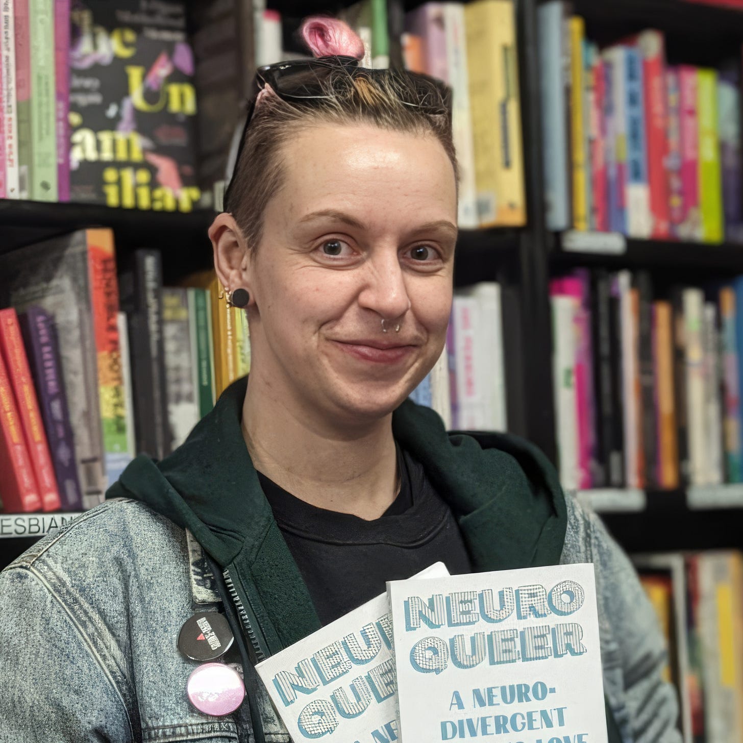 CJ DeBarra standing in a bookshop, holding two copies of Neuroqueer: A Neurodivergent Guide to Love, Sex, and Everything in Between. They wear a denim jacket over a dark T-shirt, with short hair, visible tattoos, and enamel pins on their jacket. Bookshelves filled with brightly coloured spines line the background. CJ DeBarra standing in a bookshop, holding two copies of Neuroqueer: A Neurodivergent Guide to Love, Sex, and Everything in Between. They wear a denim jacket over a dark T-shirt, with short hair, visible tattoos, and enamel pins on their jacket. Bookshelves filled with brightly coloured spines line the background.