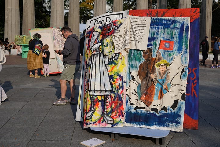 A woman holding a white bag examines a brightly colored painted giant book. A child uses a marker to add to a book made out of pillows and furniture. A group of people examines a colorful giant book with painted text and images. A giant, brightly colored book stands in front of a building with columns.