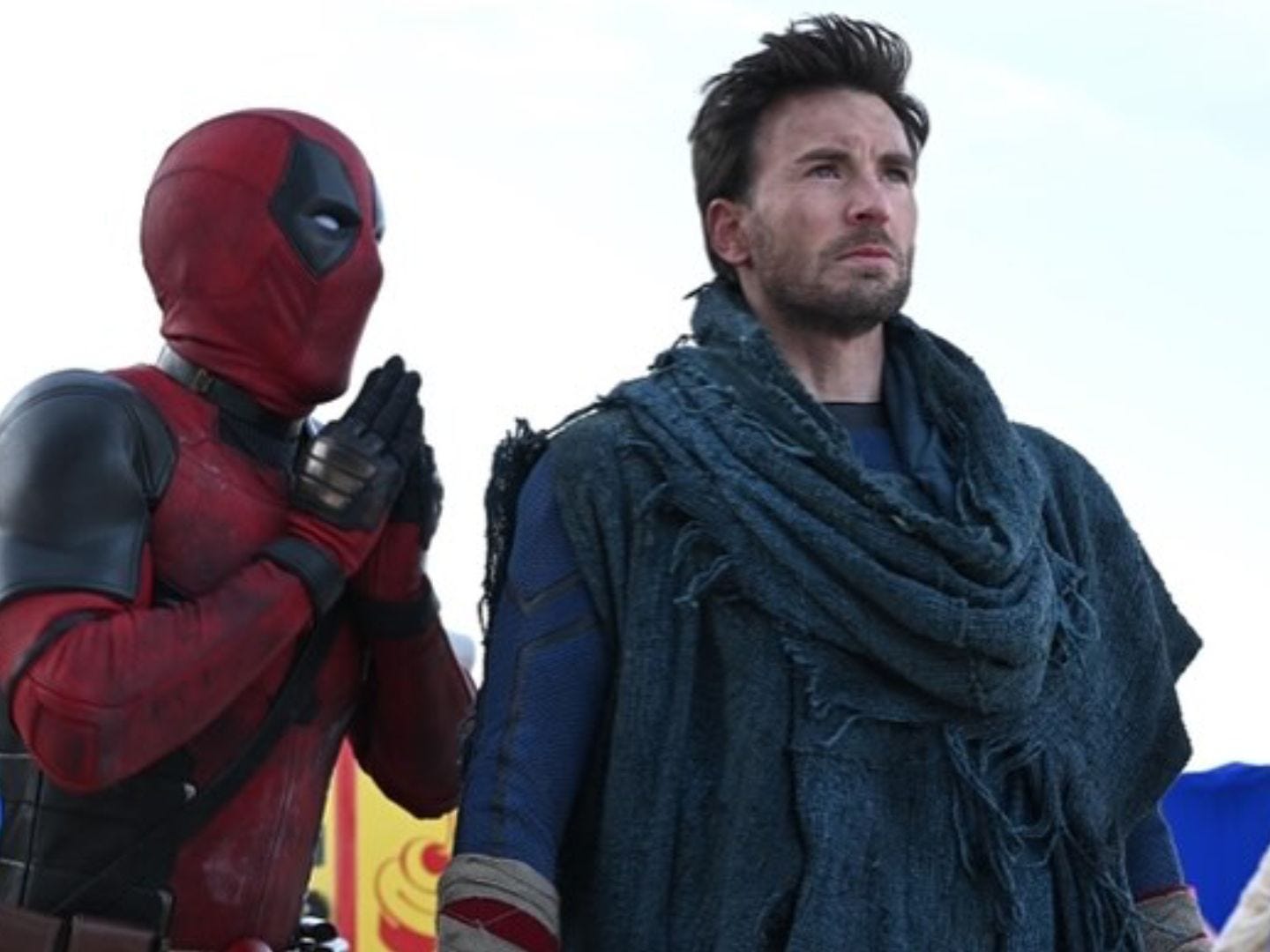 Deadpool whispers beside Chris Evans as Johnny Storm, wrapped in a blue cloak in Deadpool & Wolverine.