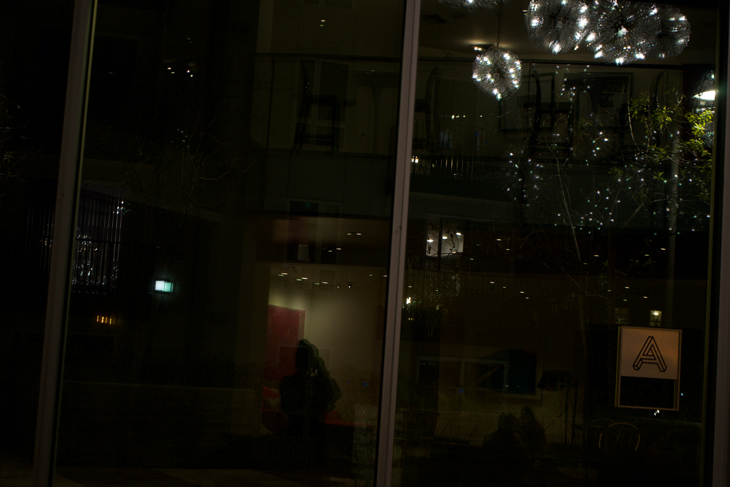 A low-lit self-portrait captured through reflective glass, where chandelier light and city reflections overlap with a silhouetted figure holding a camera. Vertical window framing divides the composition, creating depth and ambiguity between interior and exterior space.