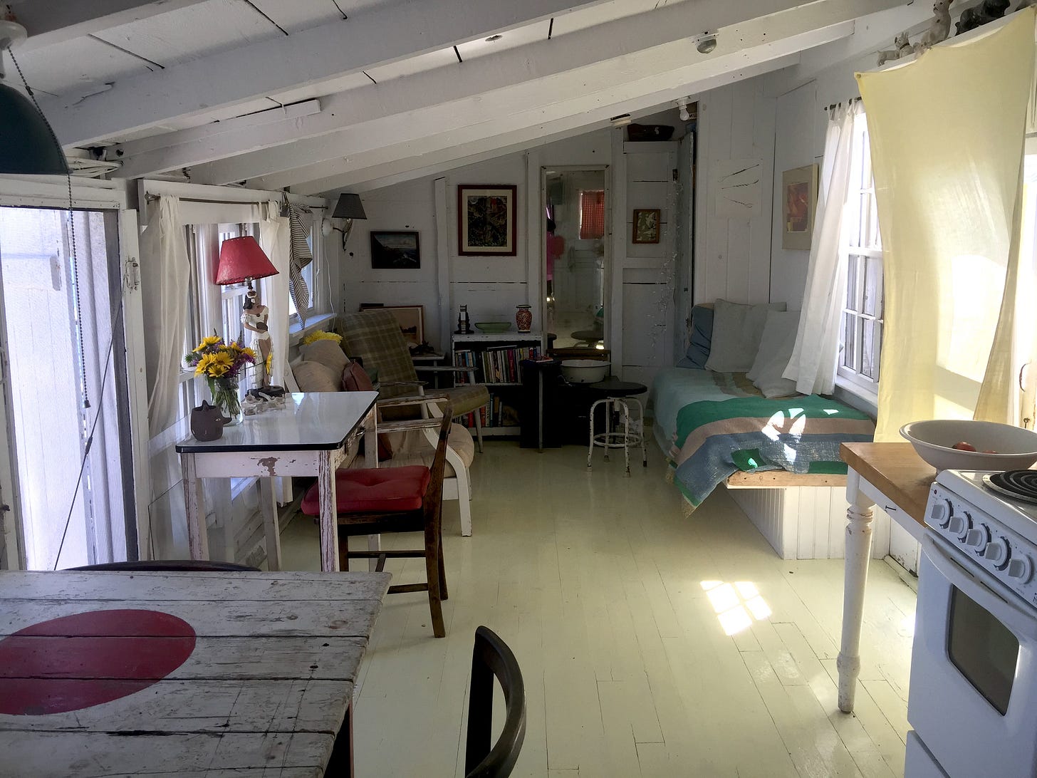 Photograph of a small shack with a low slanting roof. The interior is painted white, with a pale yellow-green floor. There are windows on both sides of the space with sunlight flooding in. You can see the edge of a stove and rough dining table, as well as a daybed, chairs, and small work desk.