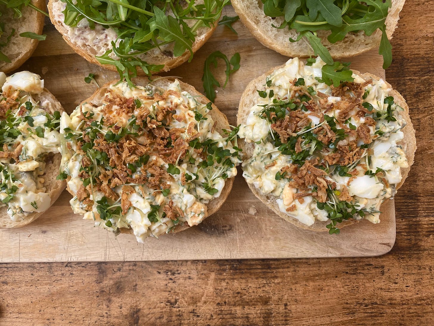 Rolls arranged on a chopping board. They are being filled with egg mayonnaise and topped with crispy bits and green salad leaves and cress.