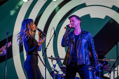 Alina Crișan & Keo doi artiști cântă împreună pe scenă, față în față, fiecare ținând câte un microfon. Artista poartă o ținută neagră, iar artistul poartă o geacă din piele. Fundalul scenei are elemente grafice luminoase în tonuri de verde și galben, iar atmosfera este specifică unui concert live.