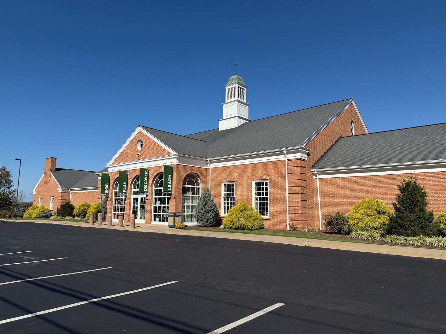 A brick building sits in front of a black paved parking lot. It has a lot of windows up front and a tiny spire. It is the World of Little League Museum