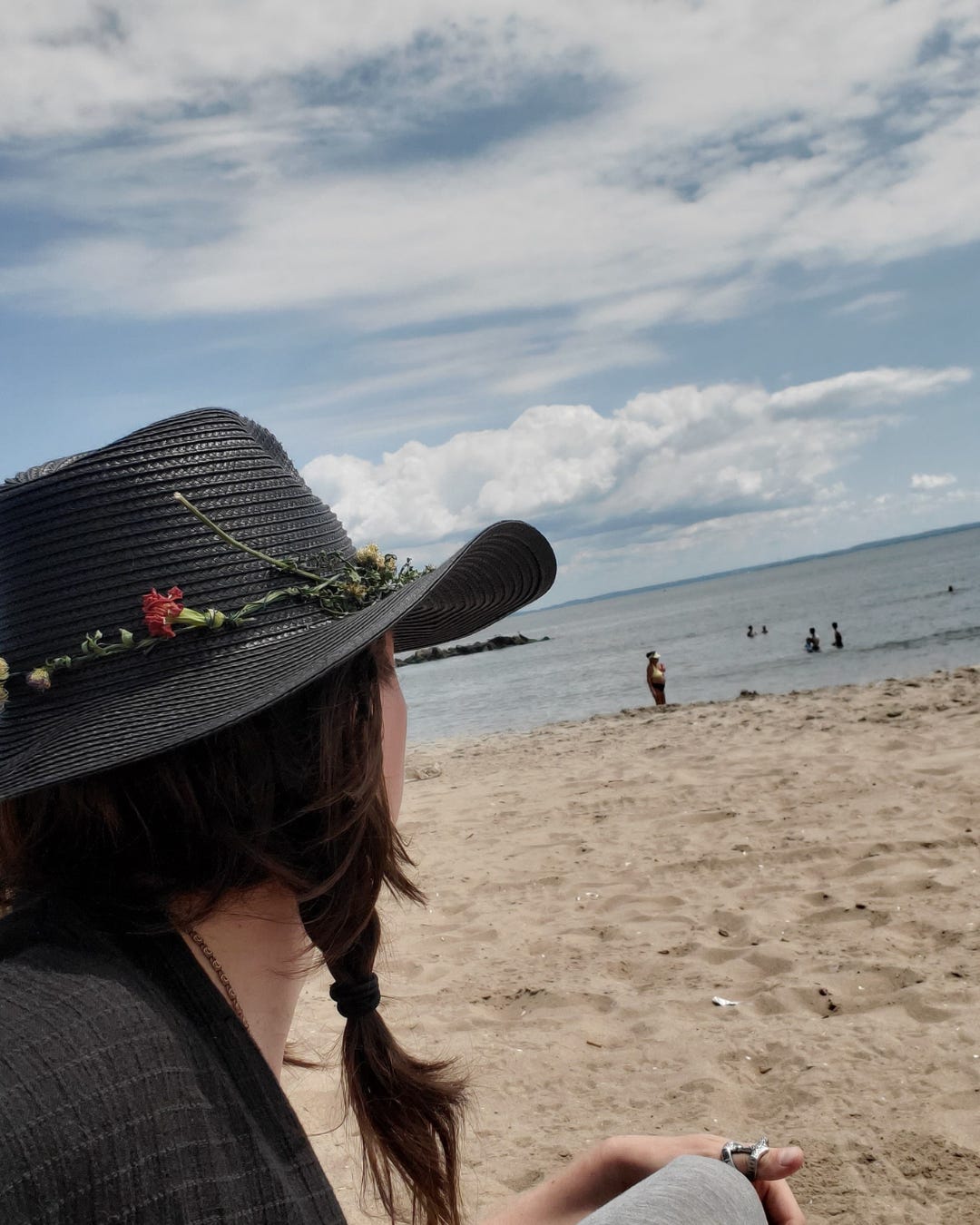 The author, crowned with a woven black cowboy hat, gazes out to the sea; the back of their head revealing only the dark braids of their hair.